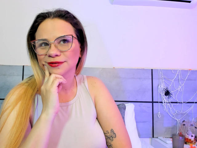 Miaamber with long blonde ponytail, glasses, red lipstick, white tank top, hand near mouth, tattoo on upper arm, cute, sitting in bedroom with purple and white lighting.