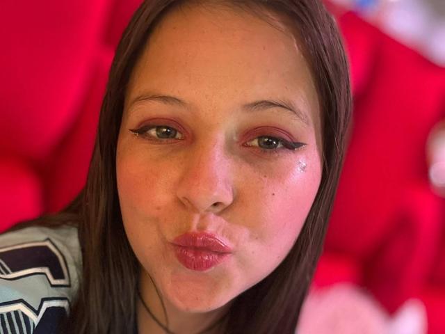 GraceAndTony has puffy pink lips, brown eyes with winged eyeliner, and freckles on her face. She's making a pouty kiss face, with her brown hair down.