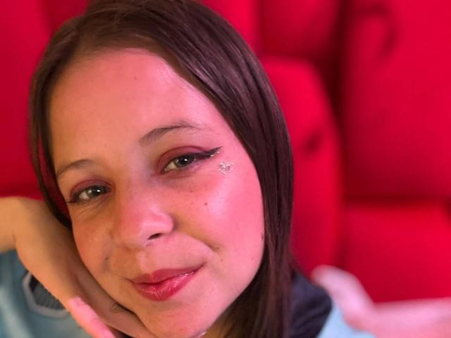 GraceAndTony has light skin, brown hair, and black eyeliner with a sparkly eye. She's resting her head on her hand, smiling with red lips, and a blue shirt.