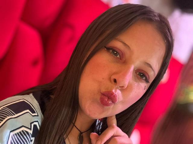 GraceAndTony, pouty lips, brown eyes, straight brown hair, wearing a black and white patterned shirt, biting her finger, red background, slightly flushed face, looking at camera.