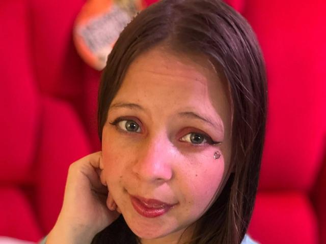 GraceAndTony has brown hair, green eyes, and a small star tattoo on her right cheek. She's smiling with red lipstick, holding her chin. Red background.