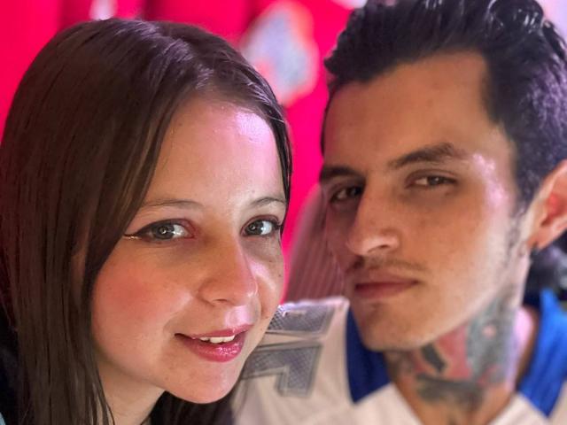 GraceAndTony has brown hair and small tits, smiling with pink lipstick. Tony has tattoos, dark hair, and a nose ring, looking at the camera with a slight smile.