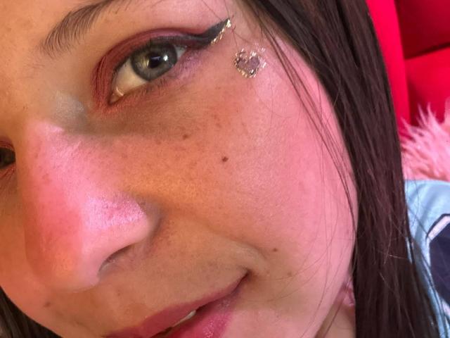 GraceAndTony's face, showing a cute, freckled nose with a small beauty mark, green eye with black eyeliner, and a gold butterfly earring, with brown hair on the right.