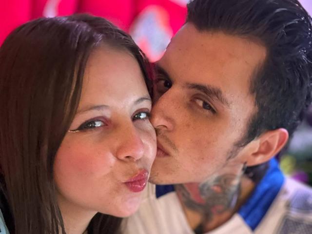 GraceAndTony with puffy red lips, dark hair, and white makeup, kissing Tony's tattooed neck. Tony has black hair, brown eyes, and a blue shirt.