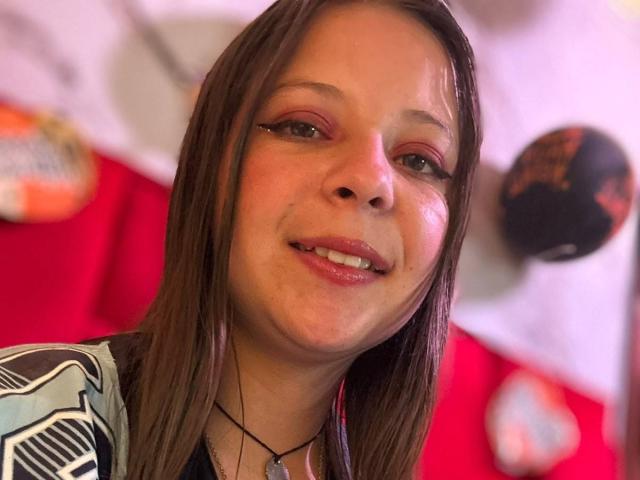 GraceAndTony has brown hair, light skin, and pinkish-red eye makeup. She's smiling with slightly parted lips, wearing a black and white patterned shirt. Blurry objects in the background.