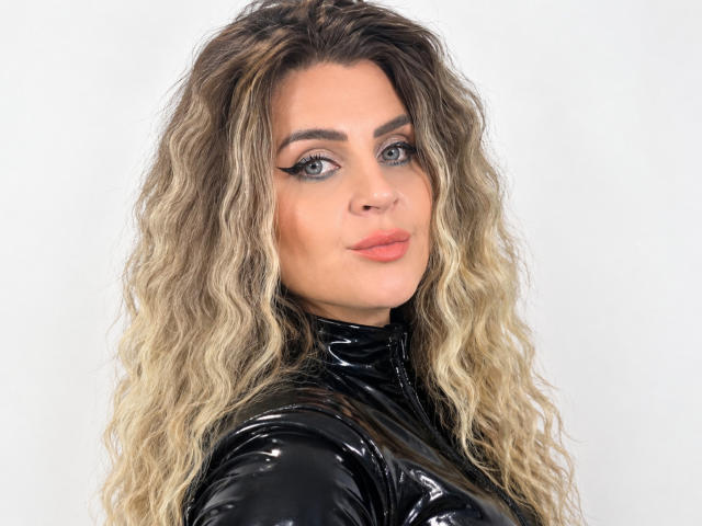 LusyBella with wavy blonde hair, blue eyes, and full lips, wearing a black glossy latex top, looking back over her shoulder with a slight smile.