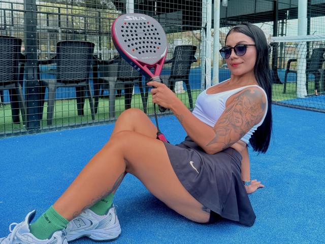 GabiHills with tan skin, black hair, tattoos, wearing sunglasses, white tank top, gray skirt, green socks, white sneakers, holding pink and black tennis racket, sitting on blue court.