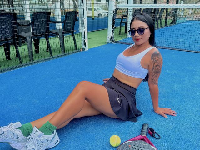 GabiHills with tan skin, black hair, and tattoos, sitting on a blue tennis court, wearing a white sports bra, black skirt, green socks, and white sneakers, with a tennis ball.