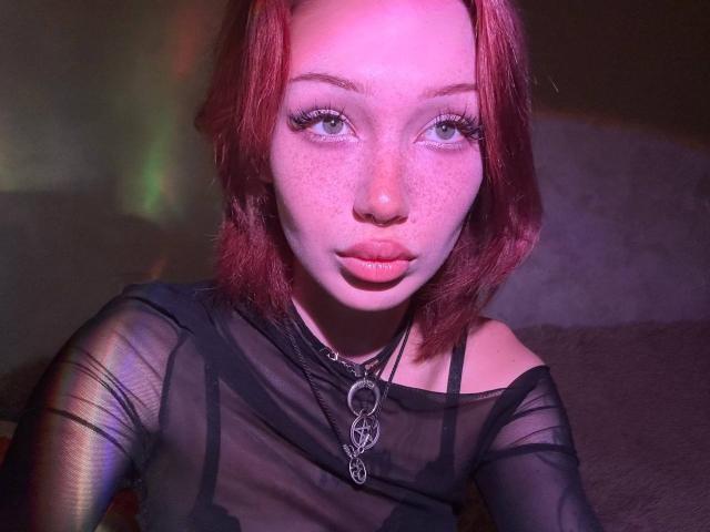 AlineGossom with red hair, freckles, and pouty lips, wearing a sheer black top, and a metal necklace with a circular pendant, looking into the camera.