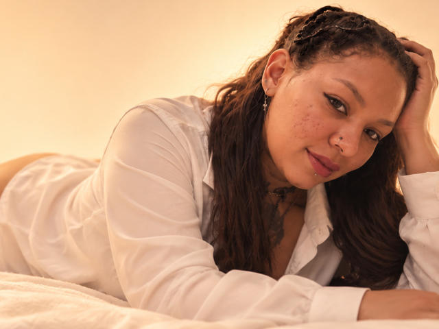 BrunnetAndWild, lying on a bed, wearing a white shirt, showing her ass, with braided hair, freckles, and a nose ring, looking at the camera with a slight smile.