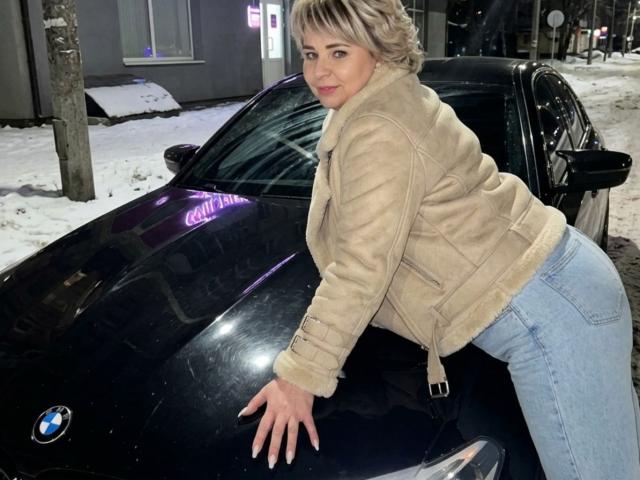 EvaCrystall leaning on a black BMW, wearing a beige suede jacket and blue jeans, showing her ass, with short blonde hair, and a slight smile in a snowy setting.