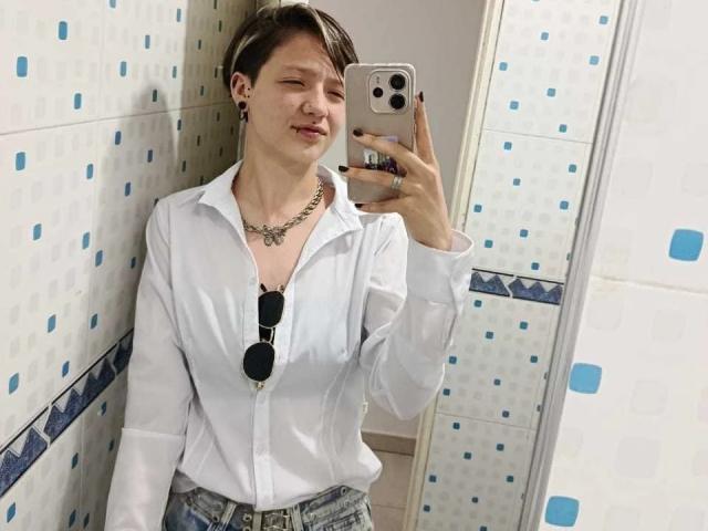 LissyLuxe with short brown hair, white button-up shirt, denim shorts, black sunglasses in pocket, necklace, and earrings, taking a mirror selfie with a slight smile.