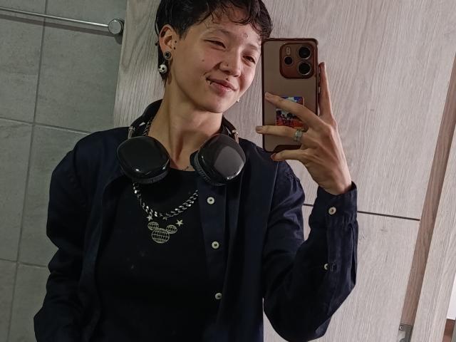 LissyLuxe, com cabelo preto curto, camisa preta e fones de ouvido, segurando um telefone, sorrindo, usando um colar, em um banheiro, olhando para o espelho..