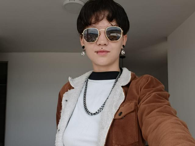 LissyLuxe, avec des cheveux noirs courts, portant des lunettes de soleil aviateur dorées, une veste en velours marron avec fourrure blanche, un t-shirt noir, un collier en chaîne et des bouchons d'oreille noirs, regardant la caméra..