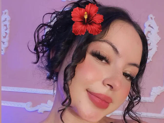 MiiaBennet with curly black hair, red hibiscus flower, winking, wearing pink lipstick, and long eyelashes, smiling softly. Her nose is small and her skin is fair.