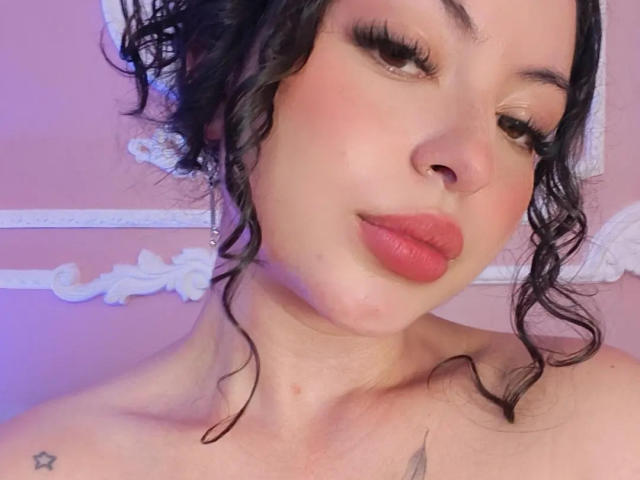 MiiaBennet has curly black hair, red lipstick, and a small star tattoo on her shoulder. She's looking down with a slight smile, showing a soft pink complexion.
