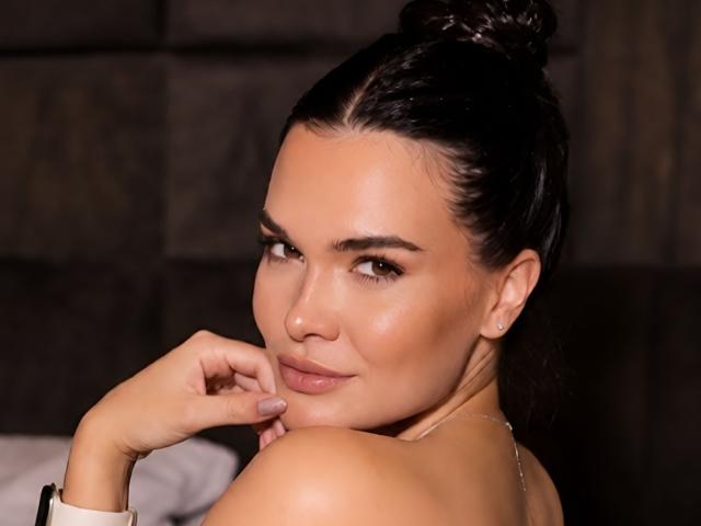 LubaMattera with dark hair in a bun, smiling at the camera, looking over her shoulder, her hand near her mouth, showing her bare shoulder and a subtle nose ring.