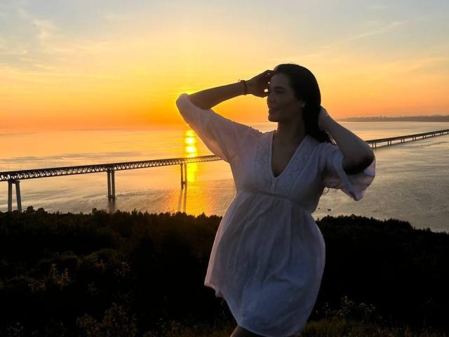 LubaMattera in a white dress, standing with hands on head, silhouetted against sunset, wearing a bracelet, water reflection, and a long bridge in the background.
