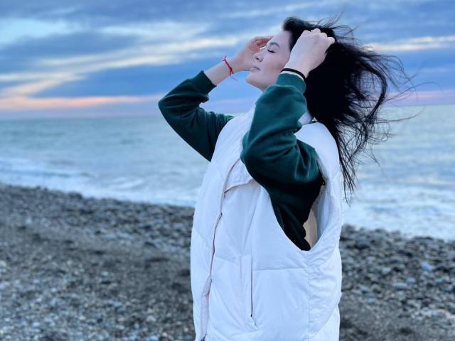 LubaMattera with dark hair, green sleeves, and white vest, stands on pebbly beach, hands in hair, eyes closed, ocean and sunset in background, wearing red bracelet.