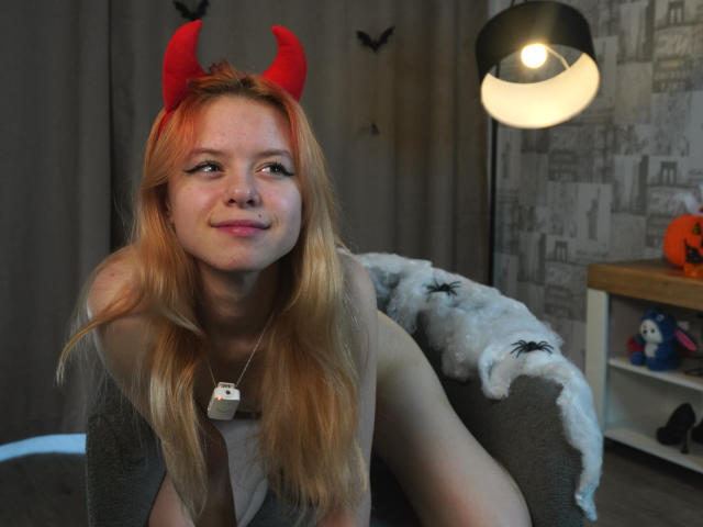 JoiMazey with red devil horns, smiling, has long blonde hair, light skin, small breasts, wearing a necklace, in a room with a black lamp and gray-patterned wallpaper.