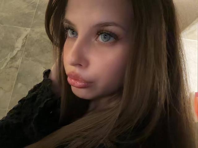 El rostro de TonieZeigler, mostrando ojos azules, labios rosados brillantes y cabello castaño largo, vestida con una blusa negra de encaje, en un selfie..