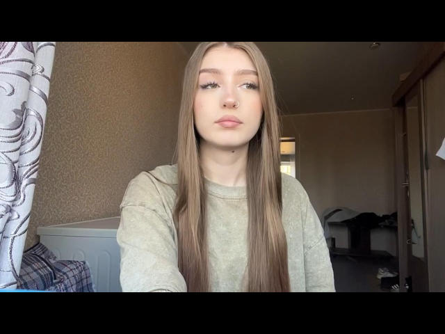 EleanoraMirabal has long, straight brown hair, a nose ring, and is wearing a grey sweatshirt, looking at the camera with a slight pout, in a softly lit room.