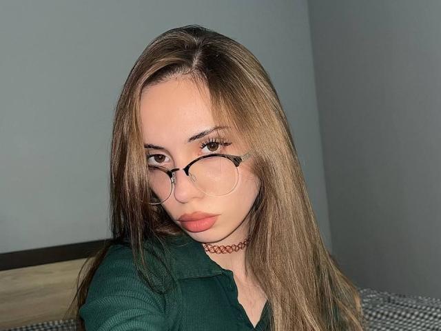 JudyPalas, com cabelos castanhos longos, óculos, camisa verde, colar vermelho e lábios rosados, olhando sedutivamente para a câmera. Seios não visíveis. Suas nádegas não estão no enquadramento..