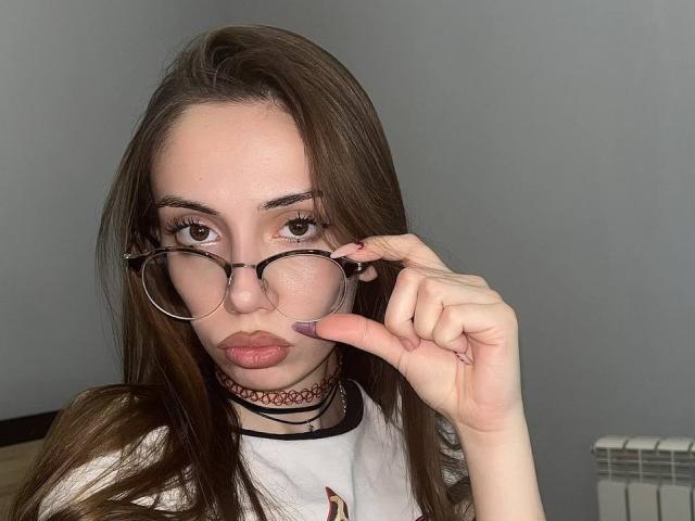 JudyPalas, met lang bruin haar, die ronde brillen draagt, een choker en een witte T-shirt, duwt haar bril op met een roze gelakte vinger en laat daarbij haar volle lippen zien..