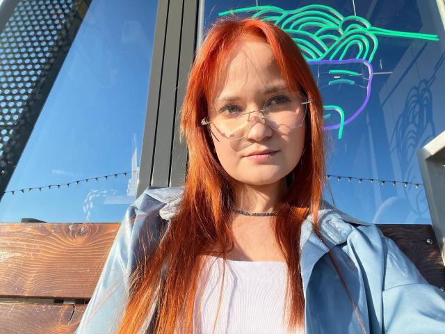 DarleneModest with red hair, glasses, and a light blue jacket, wearing a white shirt and black choker, standing in front of a neon sign.