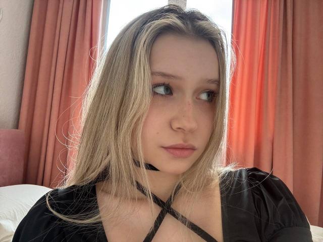 RaeleneRaya with blonde hair, black choker, and black dress, gazing sideways with slight pout, small nose, light skin, soft pink lips, and subtle makeup. Bedroom setting.