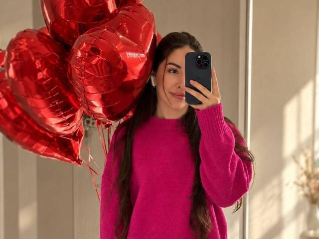 AquaLady with long brown hair, pink sweater, holding red heart balloons, taking a selfie with a black phone, smiling, natural light, soft shadows, indoors.