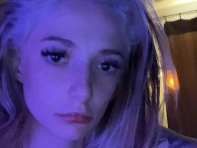 PamelaAughenbaugh with blue-tinted face, freckles, long purple hair, and thick eyelashes, looking slightly downward with a neutral expression. Her tits are not visible.