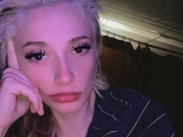 PamelaAughenbaugh has blonde hair, full red lips, and is looking at the camera with one hand on her cheek. She's wearing a striped shirt, and the lighting is purple.