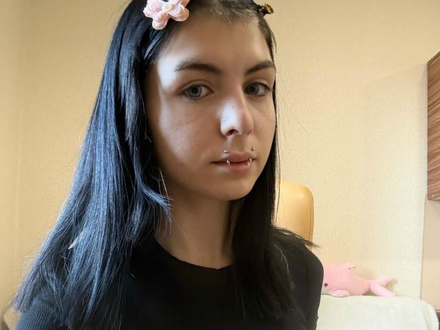 NobukoGraue has black hair, a pink flower headband, a nose ring, and a lip piercing. She's wearing a black shirt, with light skin and green eyes.