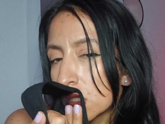 ZarayFeet with black hair, slightly sweaty face, holding black fabric in mouth, pink nail polish, and small earrings, looking down with half-closed eyes.