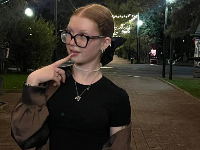 DeedraDomann, with glasses, red hair in a bun, black sheer sleeves, black top, finger to lips, necklace, standing on a brick path at night, looking cute.