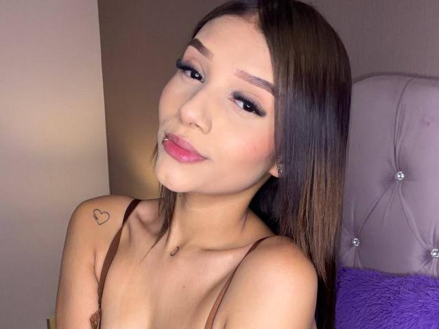 SaraMillerOne with straight brown hair, pink lipstick, and a heart tattoo on her shoulder, looking at the camera with a slight smile. She's wearing a brown top.