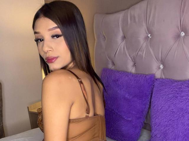 SaraMillerOne with brown hair, red lipstick, and eyeliner, wearing a brown strapless top, looking over her shoulder, sitting on a bed with purple pillows and a tufted headboard.