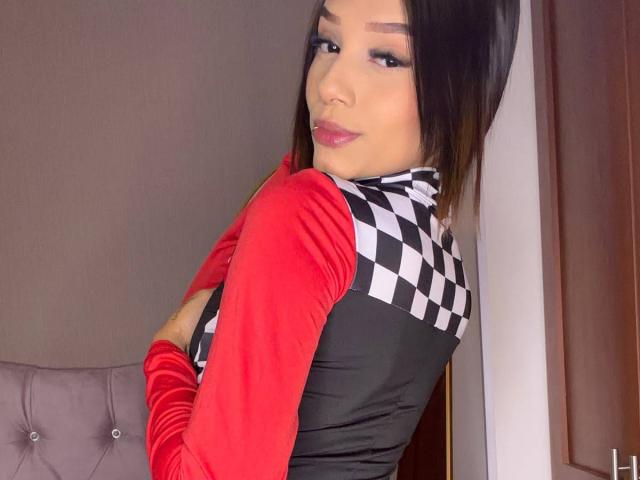 SaraMillerOne with black hair, red sleeves, and black checkered neck, crossed arms, looking over shoulder, pink lipstick, subtle makeup, sitting on gray tufted chair, smiling slightly.