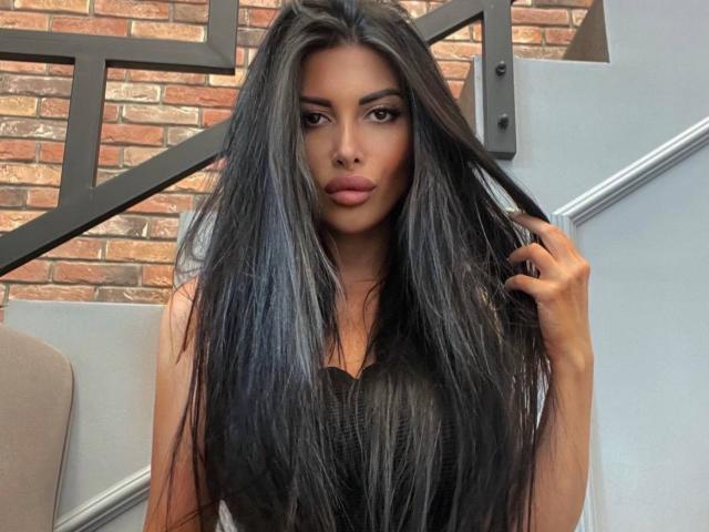 LatiaSatsky, con cabello negro largo y lacio, sosteniendo una cerradura con su mano derecha, vistiendo una blusa negra, mirando directamente a la cámara con una expresión neutra..