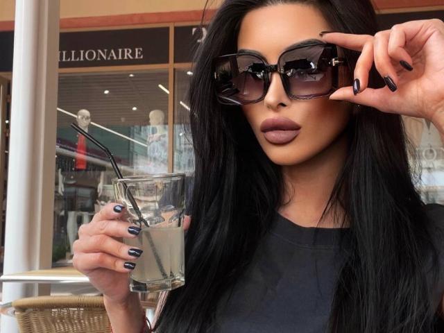 LatiaSatsky, met lang zwart haar, die zwarte zonnebril draagt, een glas met een rietje vasthoudt, zwarte nagellak heeft en een zwarte top aanheeft, ziet er cool en zelfverzekerd uit..