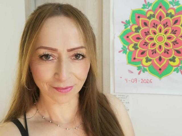 GianineLarose, con cabello castaño largo, lápiz labial rosa, maquillaje sutil, aros grandes y un collar, sonriendo a la cámara, mostrando sus senos y un colorido mandala en la pared..