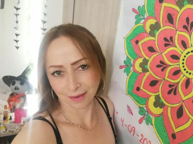 GianineLarose, con cabello castaño, aros grandes, un top negro con tirantes, maquillaje discreto y un collar, está de pie junto a un colorido dibujo de mandala, sonriendo suavemente. Se pueden ver parcialmente sus senos..