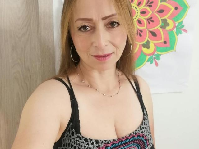 GianineLarose, con cabello castaño claro, aros grandes y una blusa negra y rosa con estampado, que muestra sus senos. Está sonriendo, lleva un collar de plata y un maquillaje sutil..