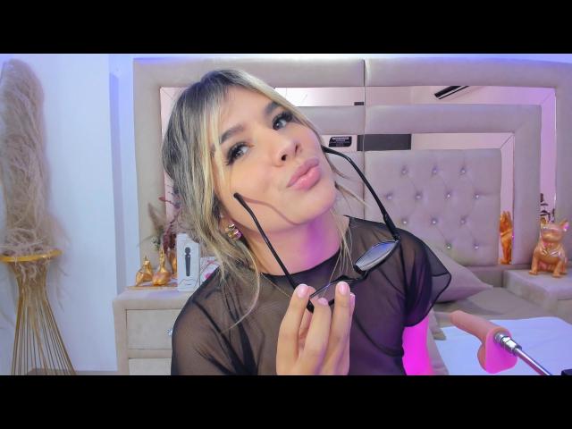 SashaPolet with blonde hair, pouty lips, and black sheer top, holding eyepiece, wearing earbuds, in a brightly lit room with pink and purple lights.