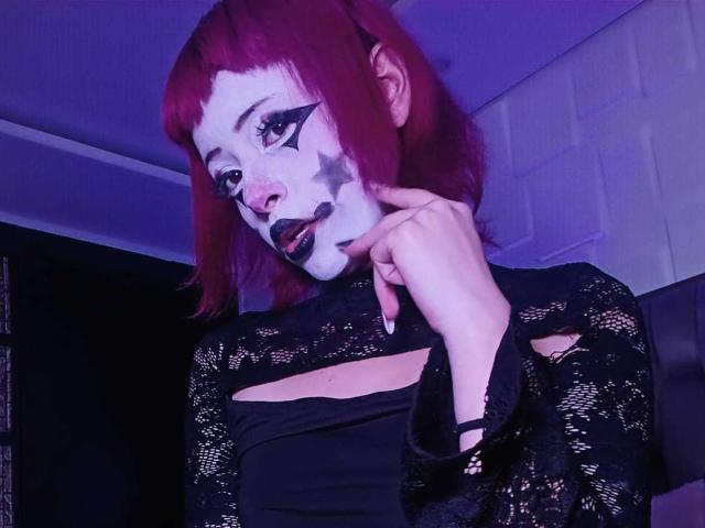 ViolettClay with red hair, white face paint, black star on cheek, black lipstick, finger on chin, wearing black lace top, cut-out chest, in dim purple light.