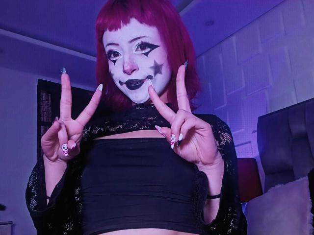 ViolettClay with red hair, white face paint, black star on cheek, black lipstick, black lace top, showing double peace signs, black nails with skull designs, in dim purple room.