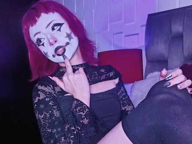 ViolettClay with red hair, white clown mask, black lace top, and black tights, holding finger to mouth, showing off her tits and black nail polish.