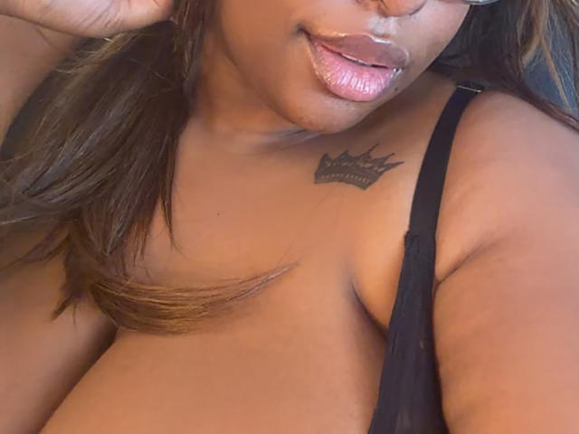 NatyLm's tan skin, glossy pink lips, and black strap on her shoulder reveal a crown tattoo between her small tits. Her brown hair falls to the side.