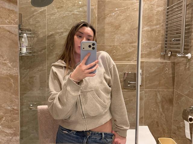 GwenOnline with straight brown hair, wearing a beige hoodie, blue jeans, and showing her bare belly, taking a mirror selfie with a grey phone in a tiled bathroom.